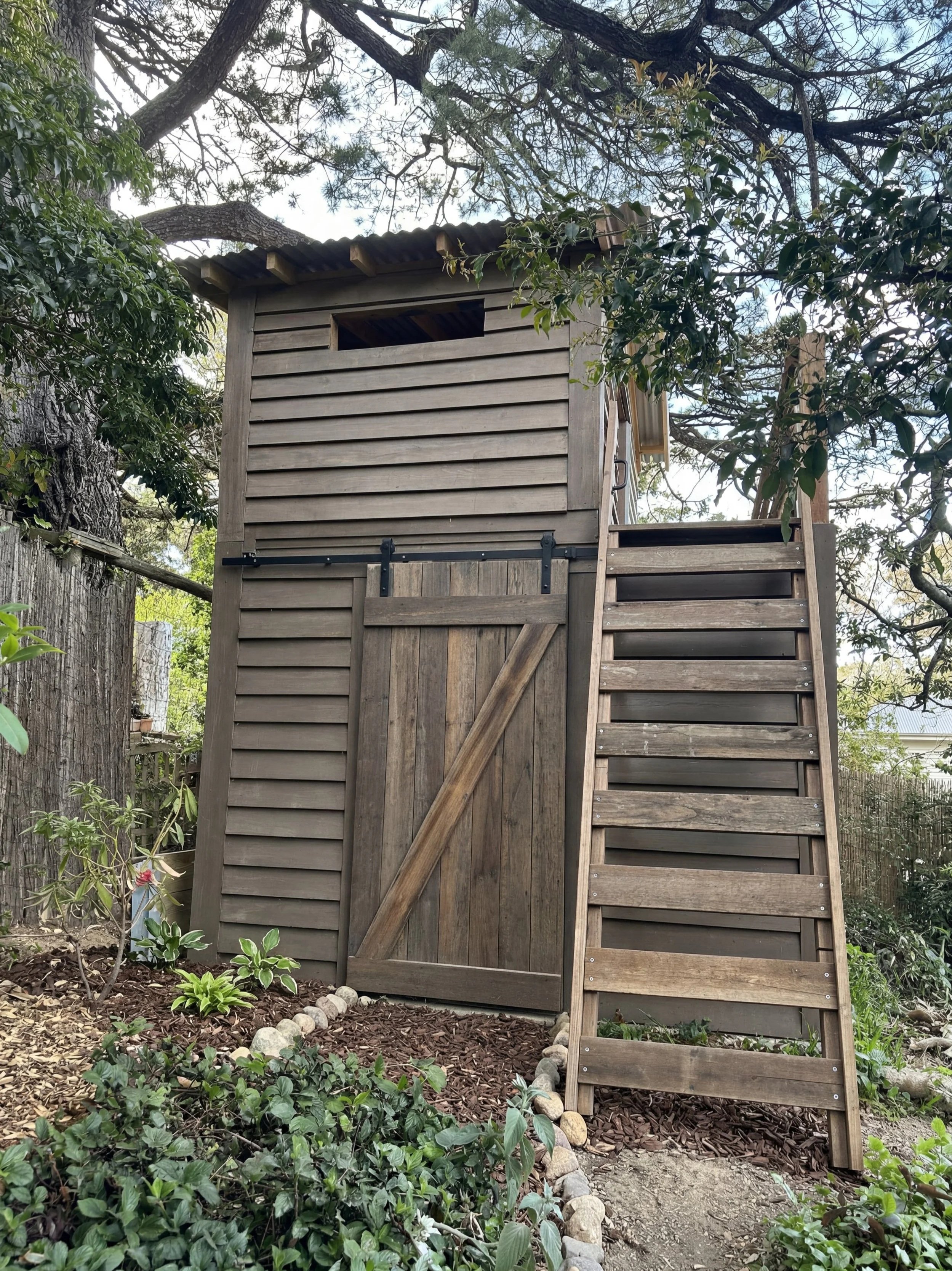 A small wooden treehouse with a staircase leading up to it, situated among trees and bushes in a garden.