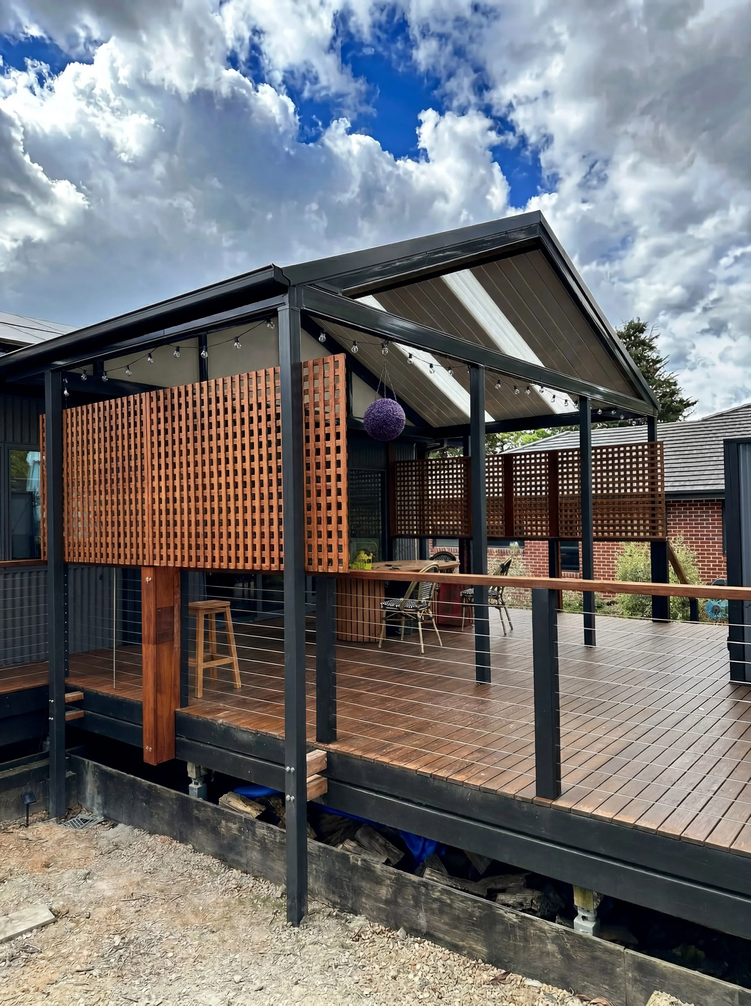 A house with a modern design featuring a raised wooden deck, dark metal frame, wooden privacy screens, string lights, and a purple paper lantern hanging from the roof, with cloudy sky overhead.