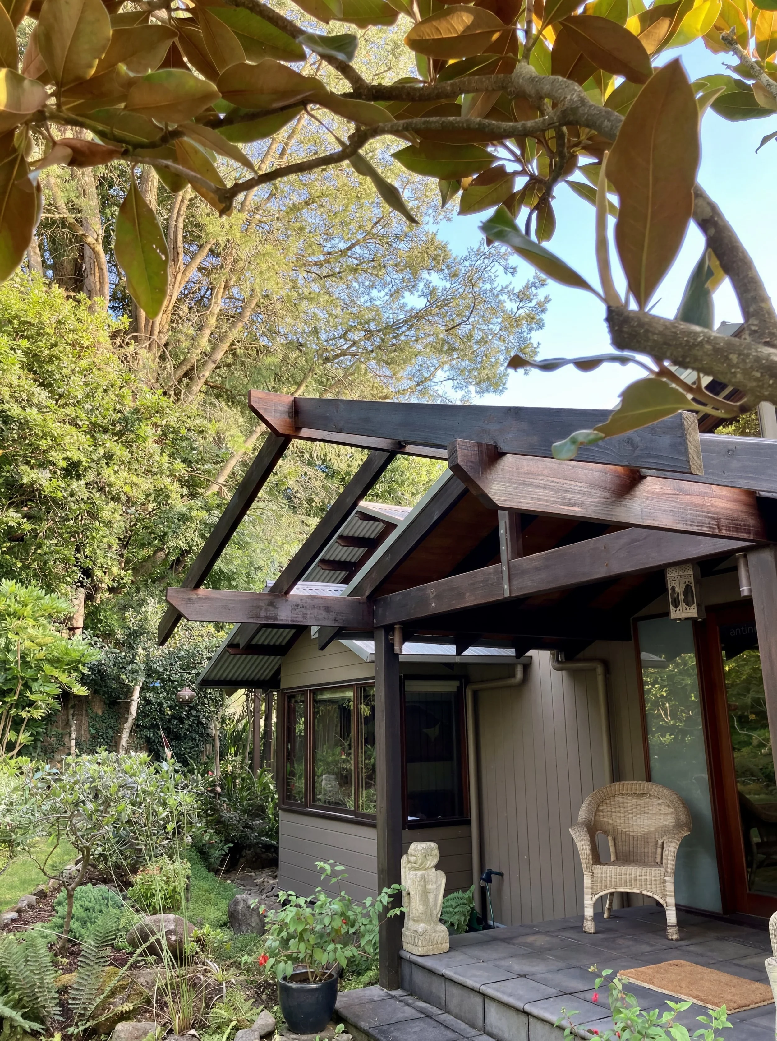Back patio of a house, with a wooden pergola, a wicker chair, and a garden full of trees and plants.