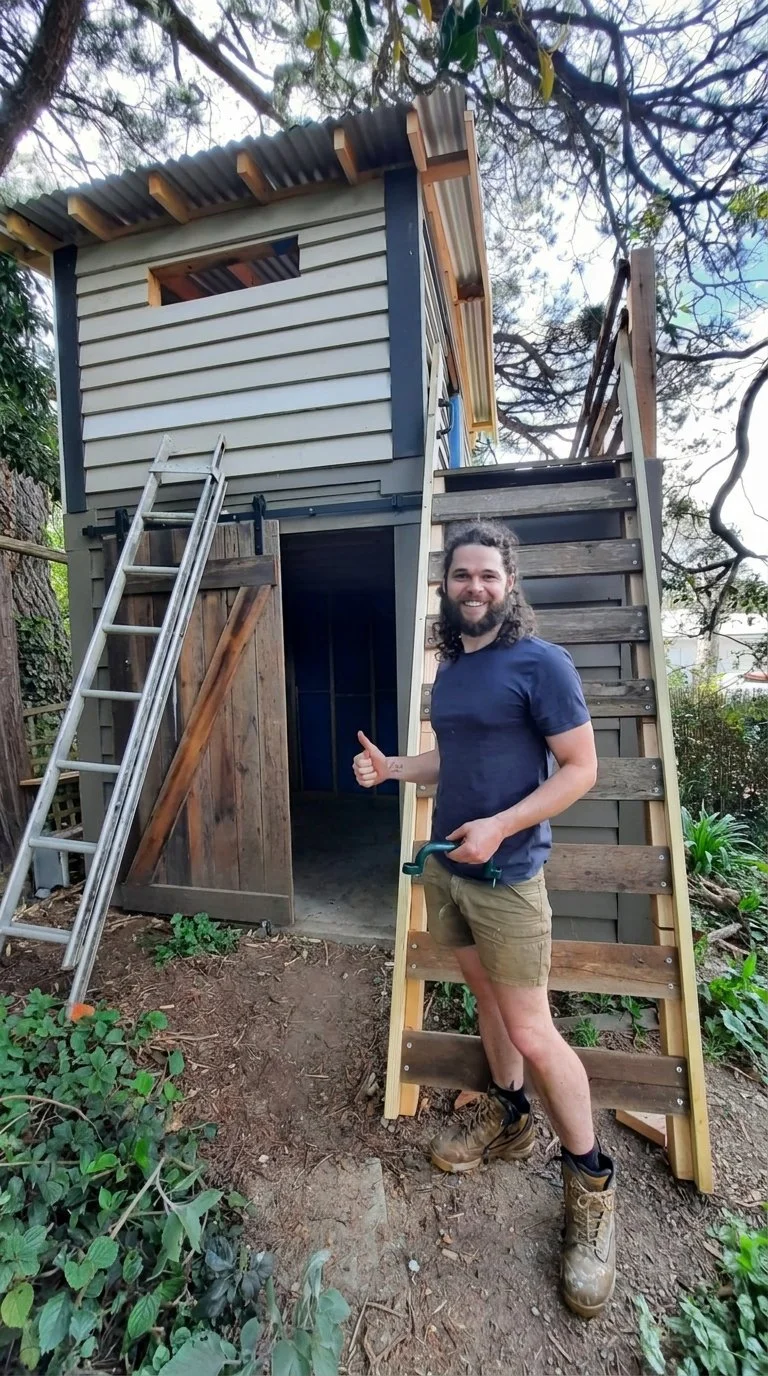 A man with long hair and a beard giving a thumbs-up while standing next to a wooden staircase leading to a small, partially built treehouse in a backyard surrounded by trees and greenery.