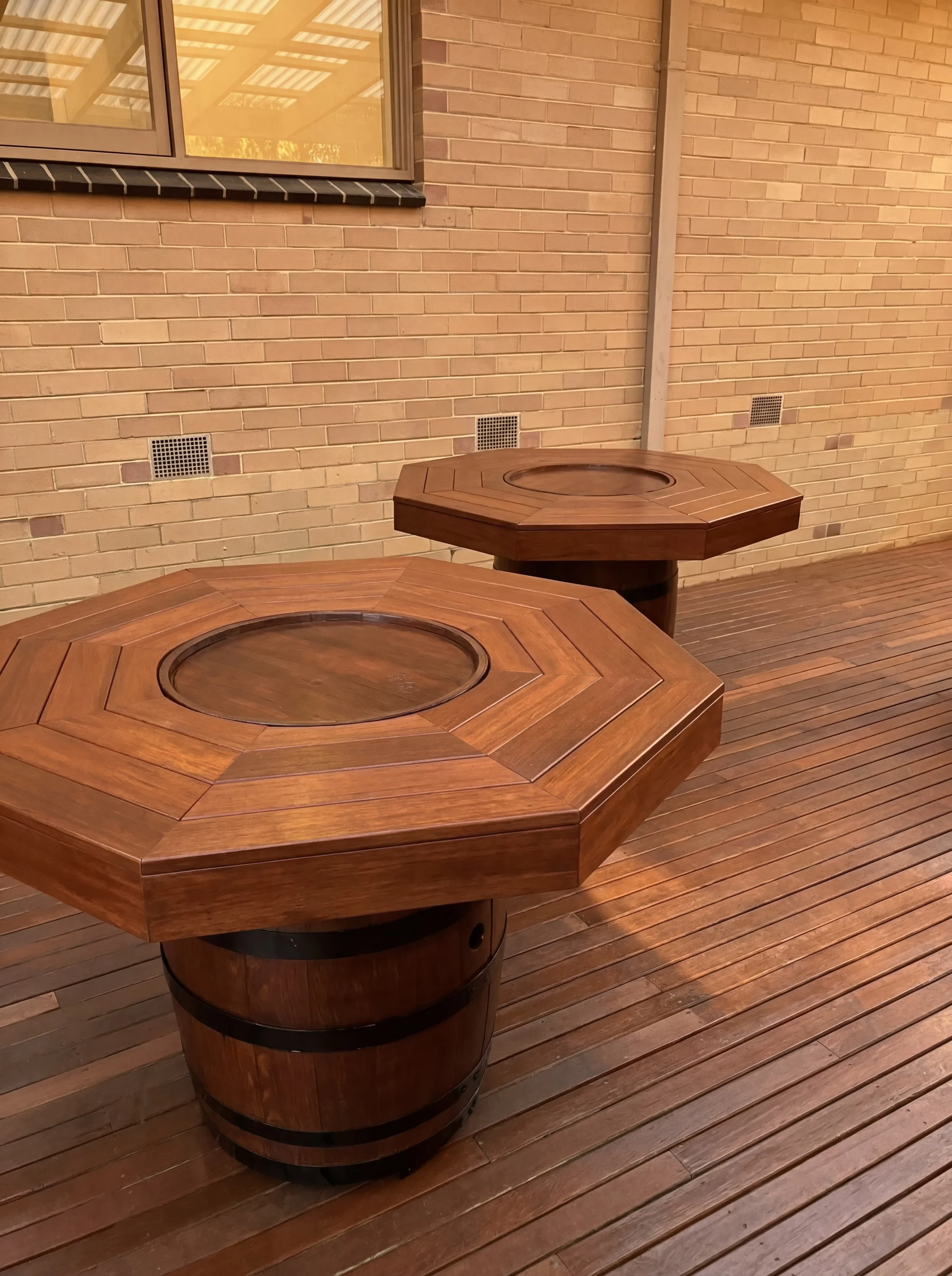 Two hexagonal wooden tables with circular inlays on top, placed on a wooden floor against a brick wall with a window.