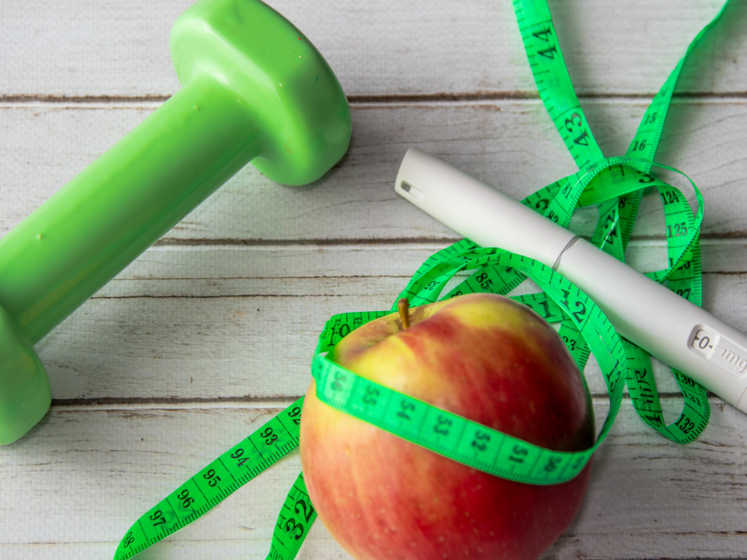 A green dumbbell, a red and yellow apple with a measuring tape around it, a white digital thermometer, and a green measuring tape on a wooden surface.