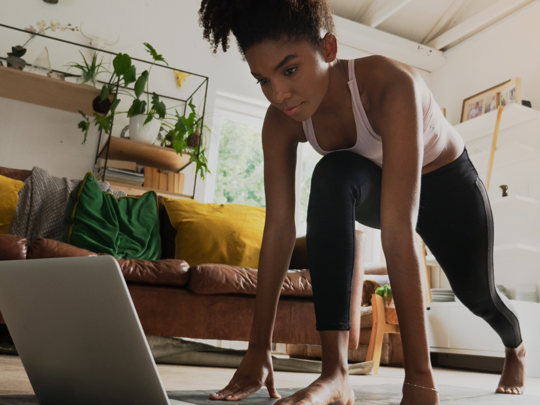 A woman in athletic wear is in a yoga pose on the floor in a living room, looking at a laptop in front of her.