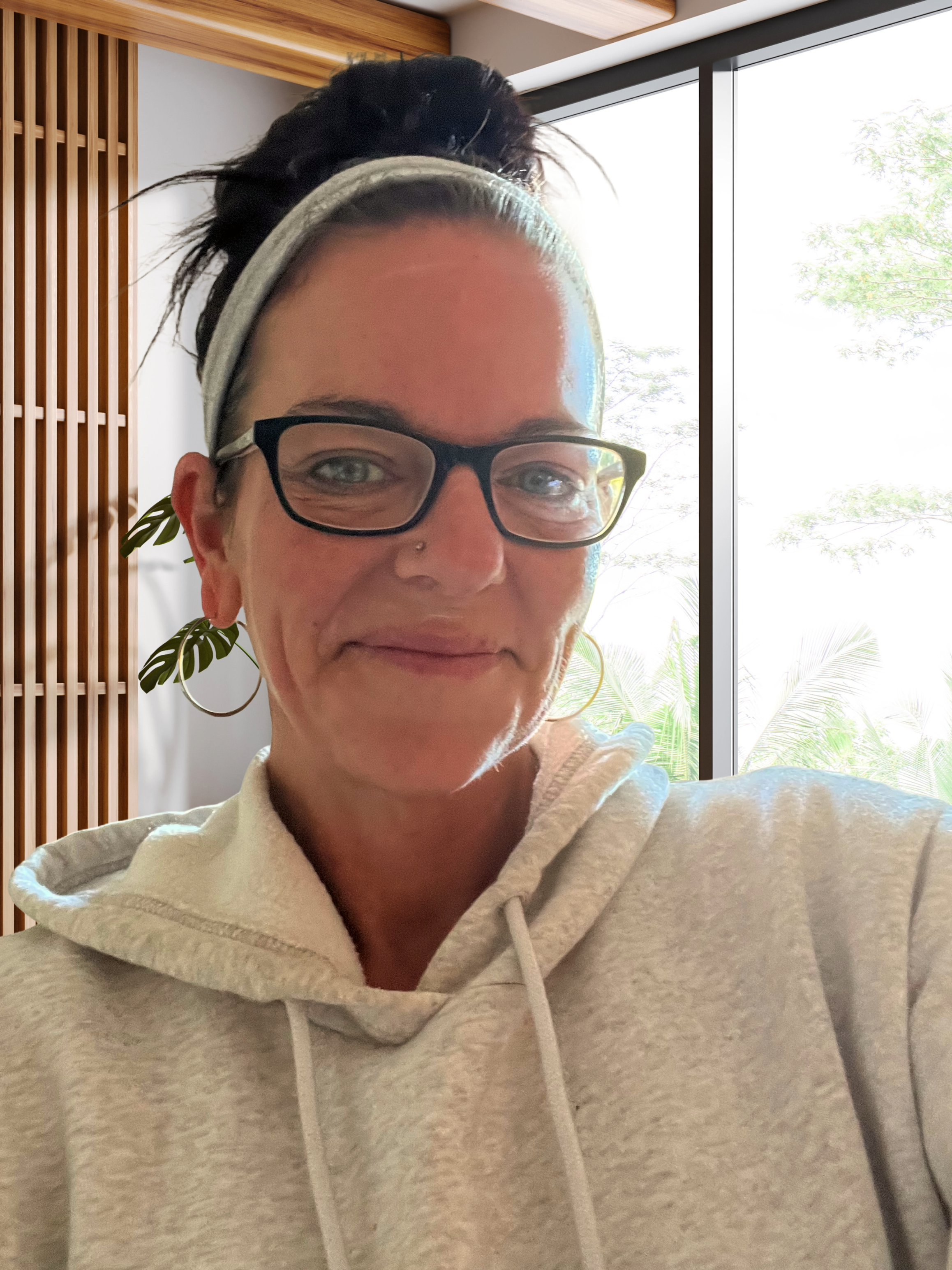 Close-up of a woman with glasses and a gray hoodie, smiling inside near a large window with greenery outside.
