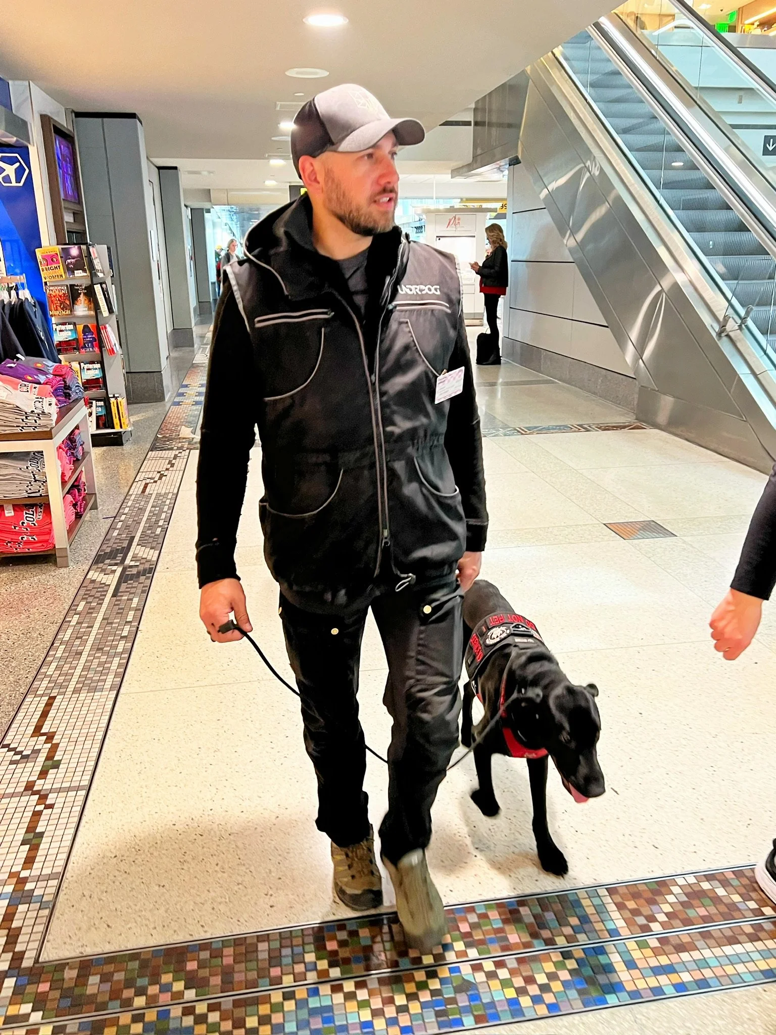 Mitch walking beside a black dog wearing a vest inside a shopping center.