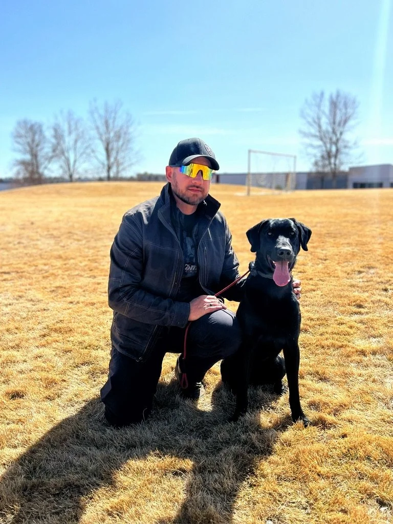 Mitch kneeling beside a black dog in a grassy field.