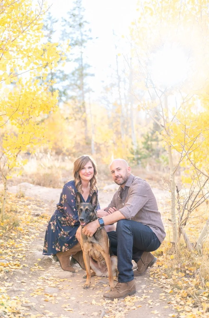 Mitch and a woman posing outdoors with a dog among autumn trees.