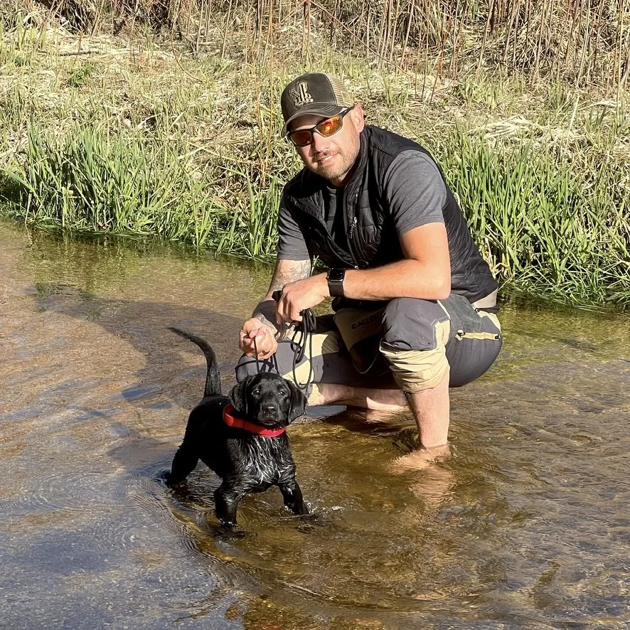 Mitch kneeling in shallow water beside a small black dog.
