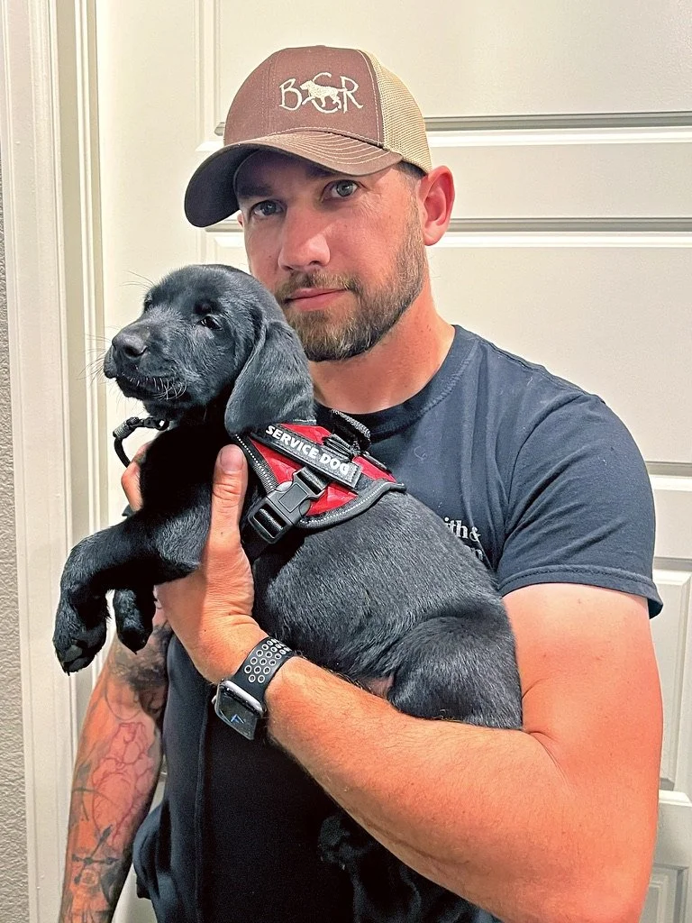 Mitch holding a black puppy wearing a red harness.