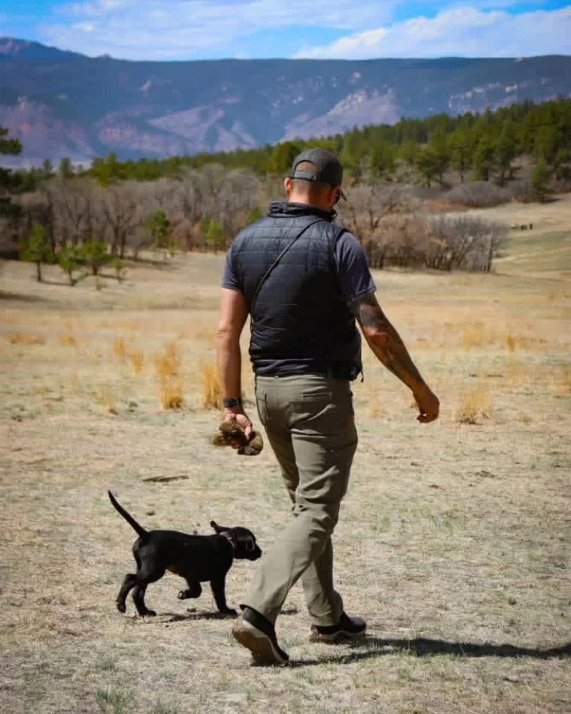 Mitch walking outdoors with a small black dog beside him.
