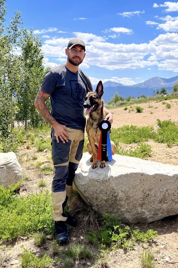 Mitch posing outdoors beside a tan and black dog with award ribbons.
