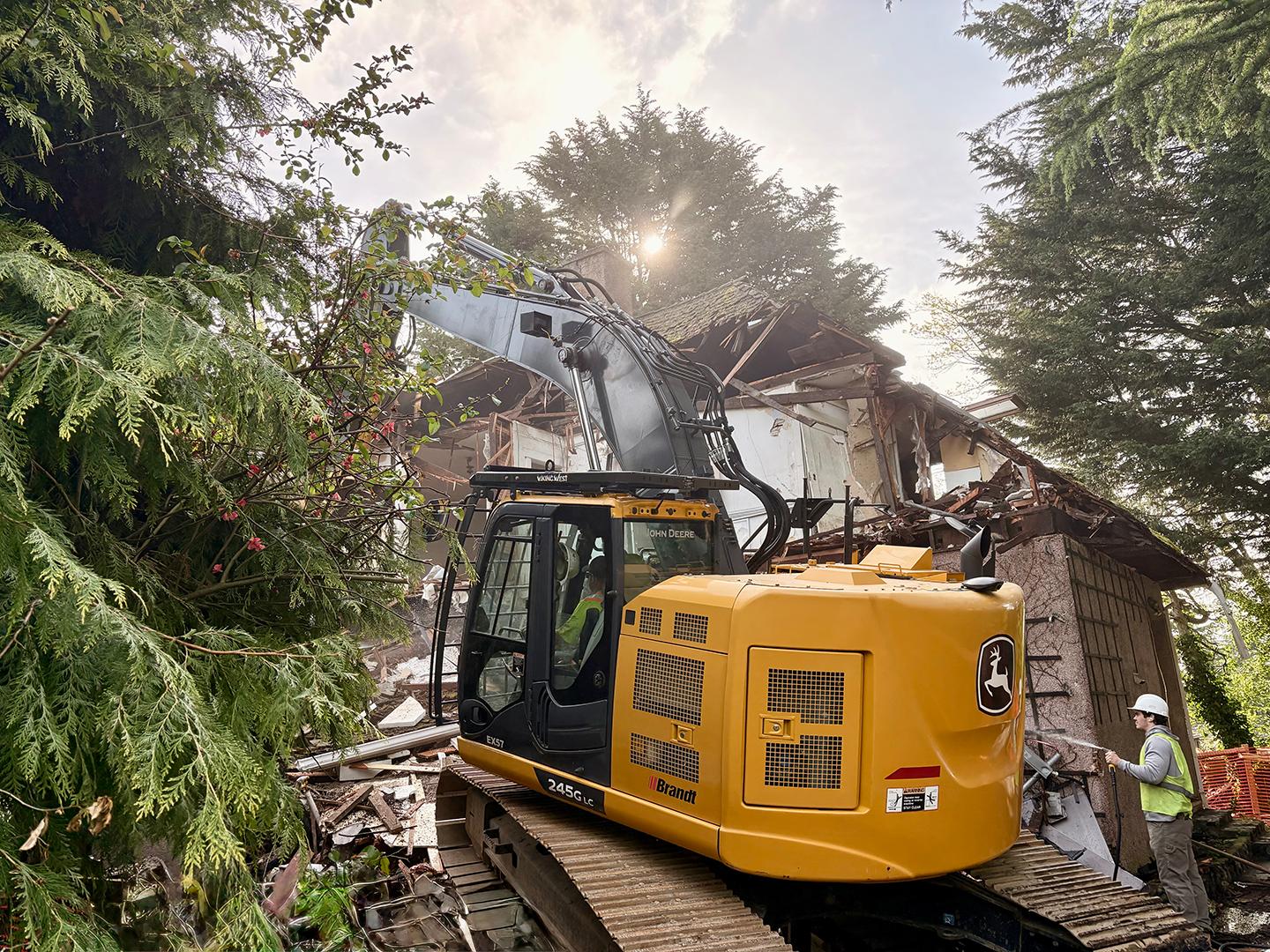 Construction worker in hard hat and safety vest using a hose to spray water near a destroyed building with an excavator clearing debris and trees surrounding the site.