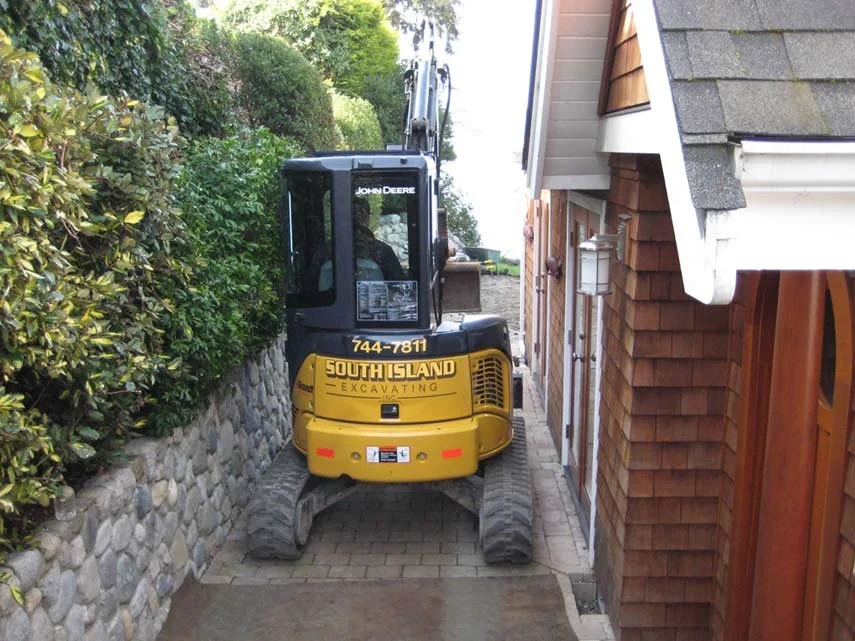 A small yellow John Deere excavator parked in a narrow alley between a stone wall with bushes and a house with wooden shingles.