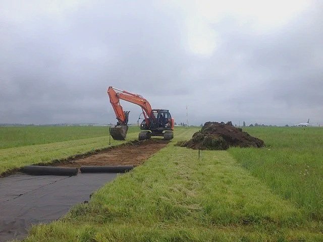 An excavator on a grassy field is digging and moving dirt, with a large pile of dirt nearby under a cloudy sky.