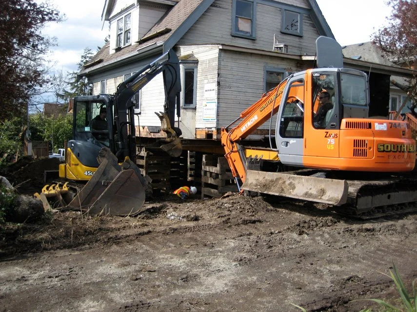 Construction site with two excavators in front of a partially demolished house, dirt and construction tools on the ground.