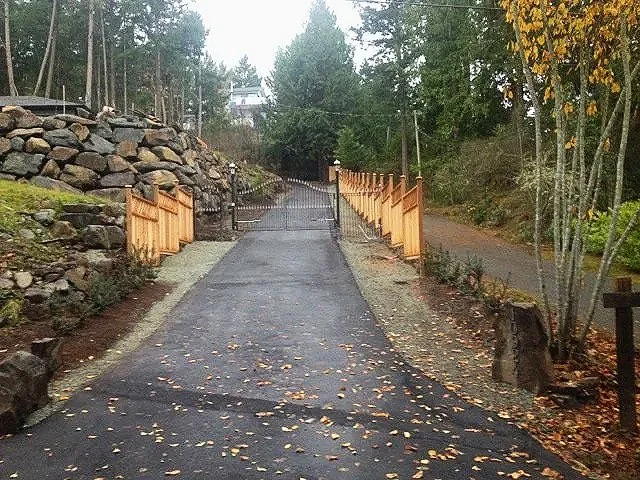 A paved path in a wooded area, with stone walls on the left and a wooden fence on the right, leading uphill through trees with autumn leaves.