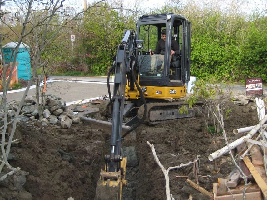 A person operating a small yellow excavator digging a trench near a sidewalk in a green area.