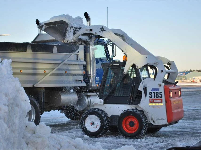 A skid-steer loader scooping snow into the bed of a truck on a snowy day.
