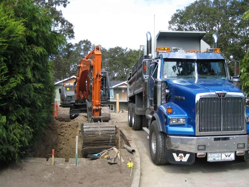 Construction site with a large blue dump truck and an orange excavator digging a trench on a residential street.