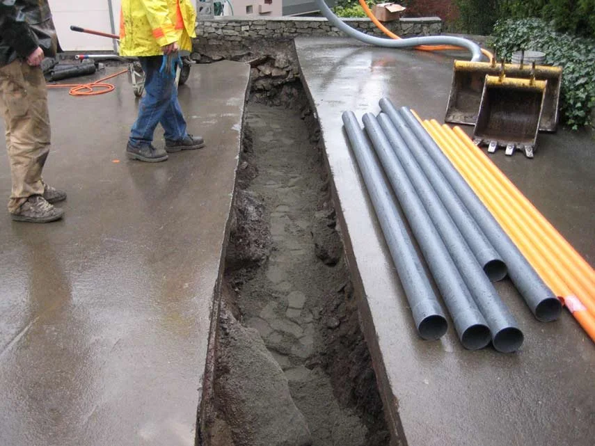 Construction workers installing underground piping in a trench on a wet sidewalk with construction tools and pipes nearby.