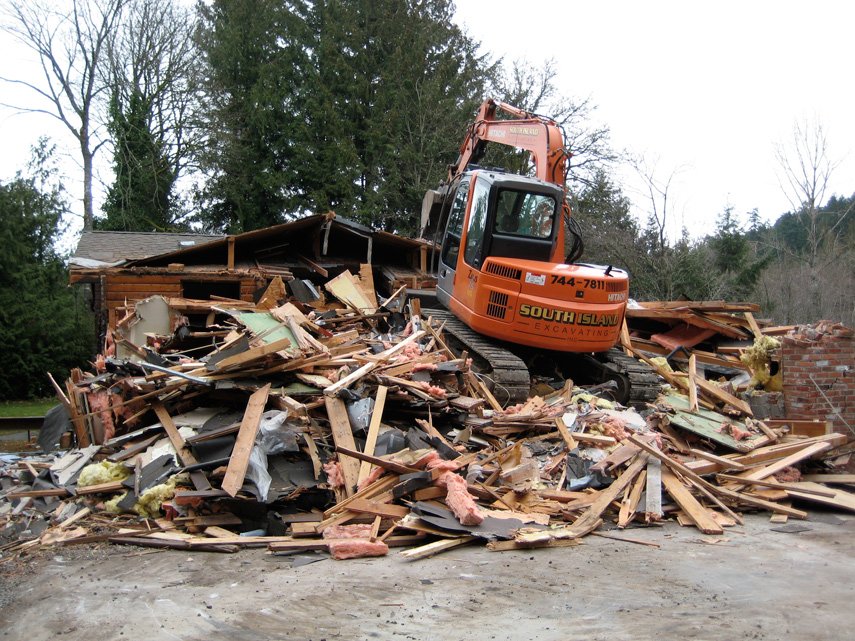 Excavator demolishing a house, debris of wood, drywall, and insulation scattered around.