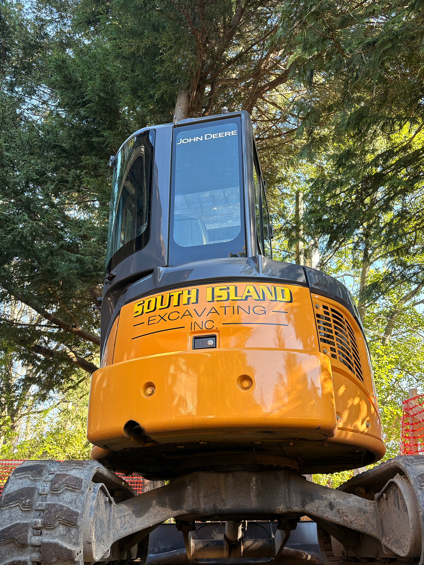 A yellow and black excavator with the words "South Island Excavating Inc" printed on its side, positioned outdoors under a large tree with green foliage.