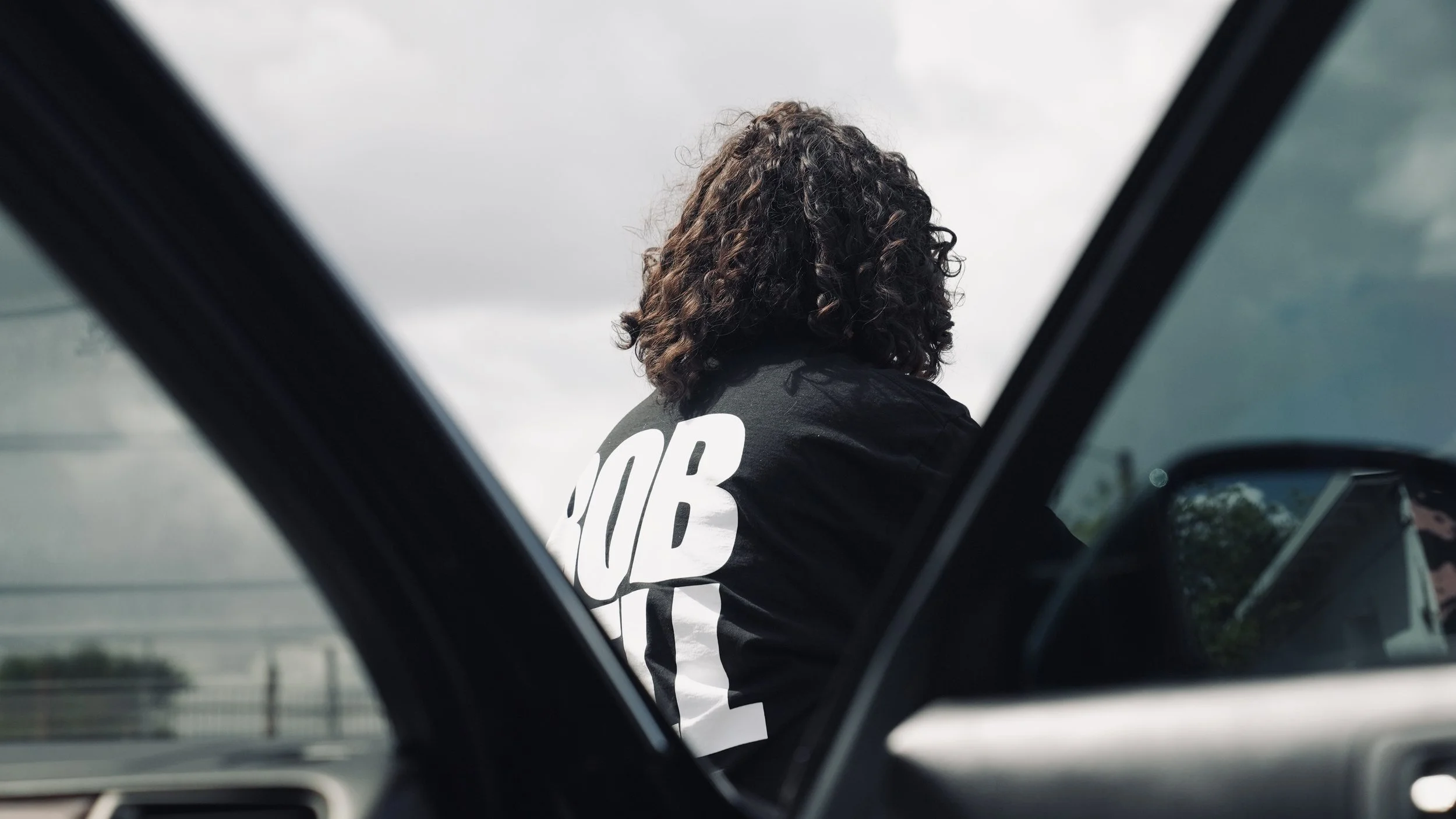 Person with curly hair wearing a black T-shirt with white text, viewed from inside a car looking out the window.