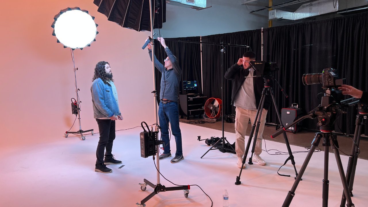 A video production crew filming a man in a studio with a plain light pink background, large studio lights, and camera equipment.