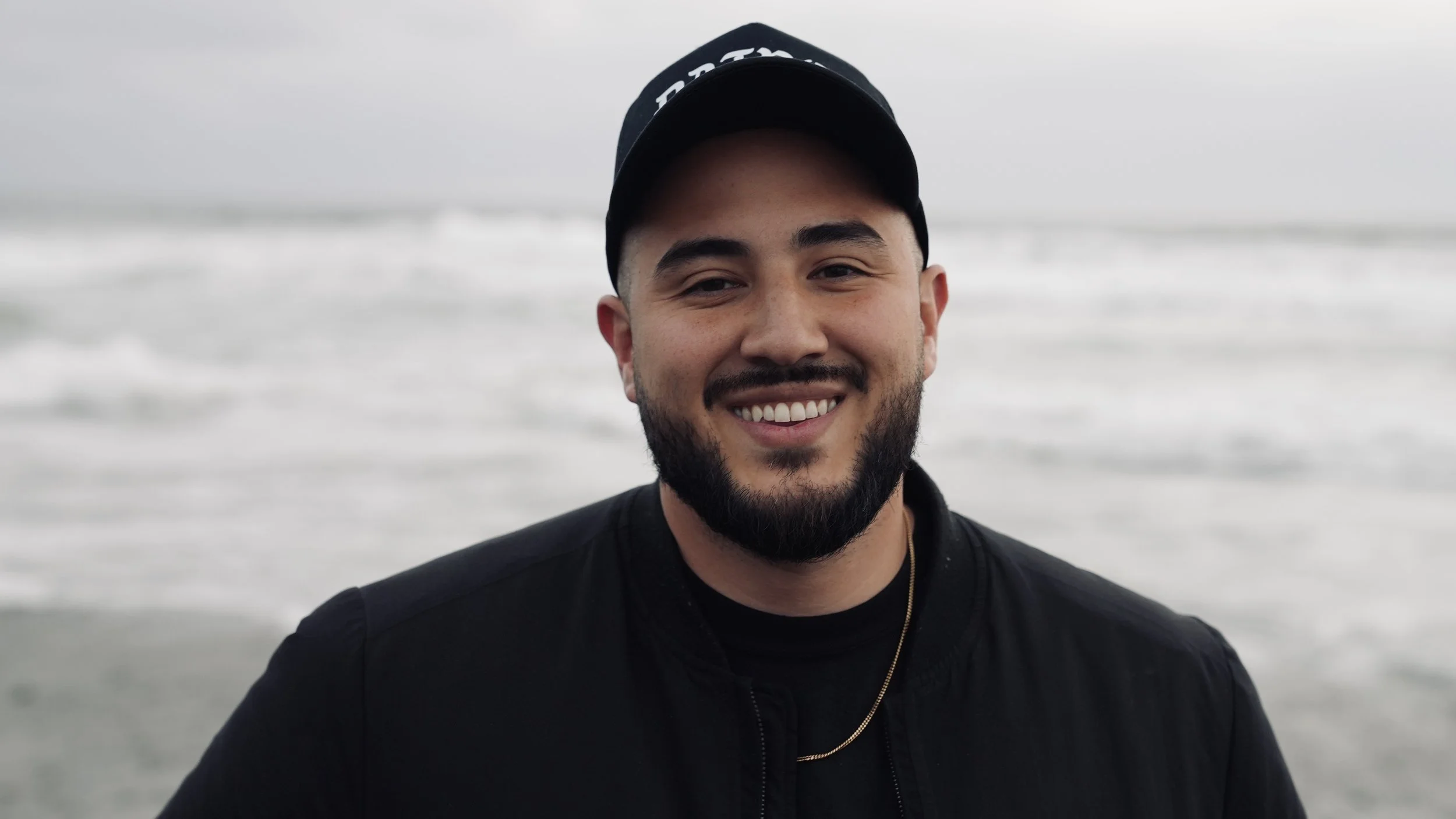 A smiling man with a beard and mustache wearing a black cap and jacket, standing on a beach with waves in the background.