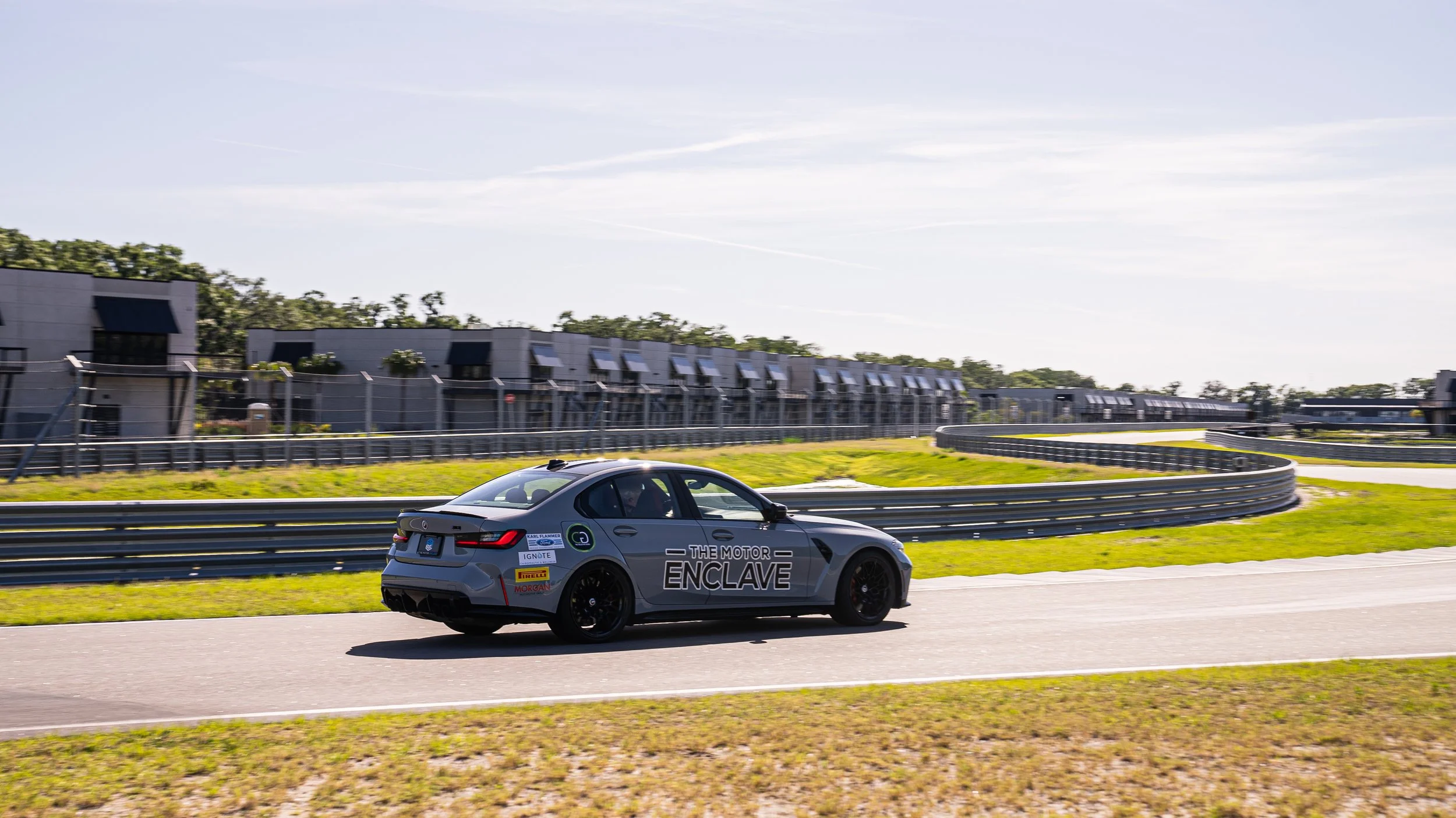 A gray race car driving on a track with buildings and trees in the background.