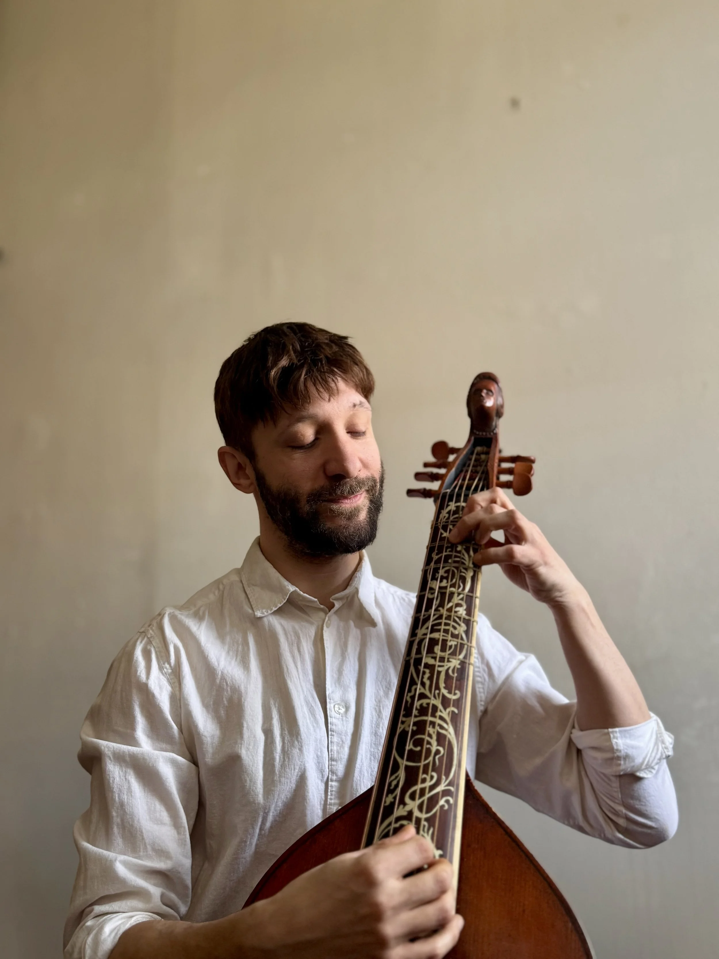 A man with closed eyes, a beard, and brown hair, wearing a white shirt, playing a stringed instrument against a plain beige wall.