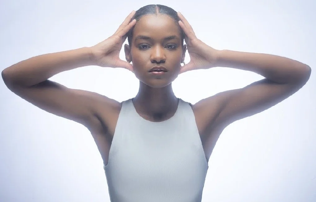 A woman with dark skin and short hair, wearing a light-colored sleeveless top, poses with her hands on her head and looks directly at the camera against a plain light background.