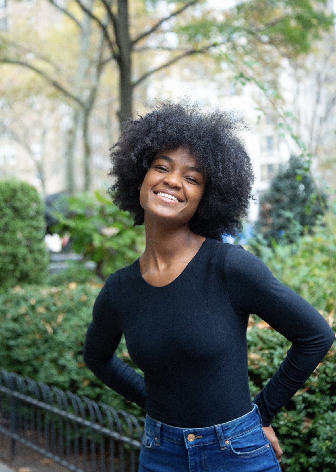 A smiling woman with natural curly black hair wearing a black long-sleeve top and blue jeans in a park with greenery and trees in the background.