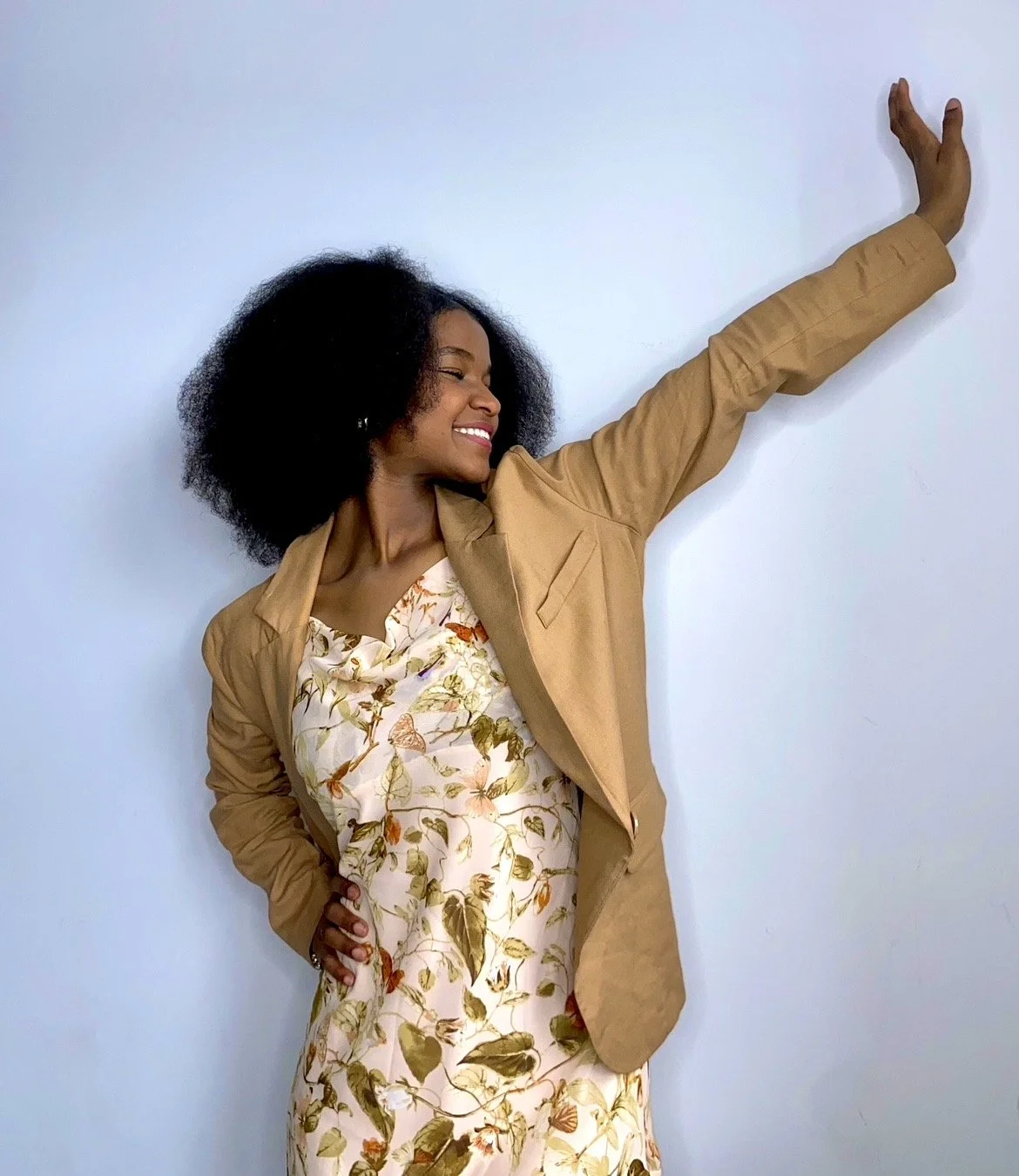 A smiling woman with curly black hair posing against a light-colored wall, wearing a floral dress and a tan blazer, with one arm raised and touching the wall.