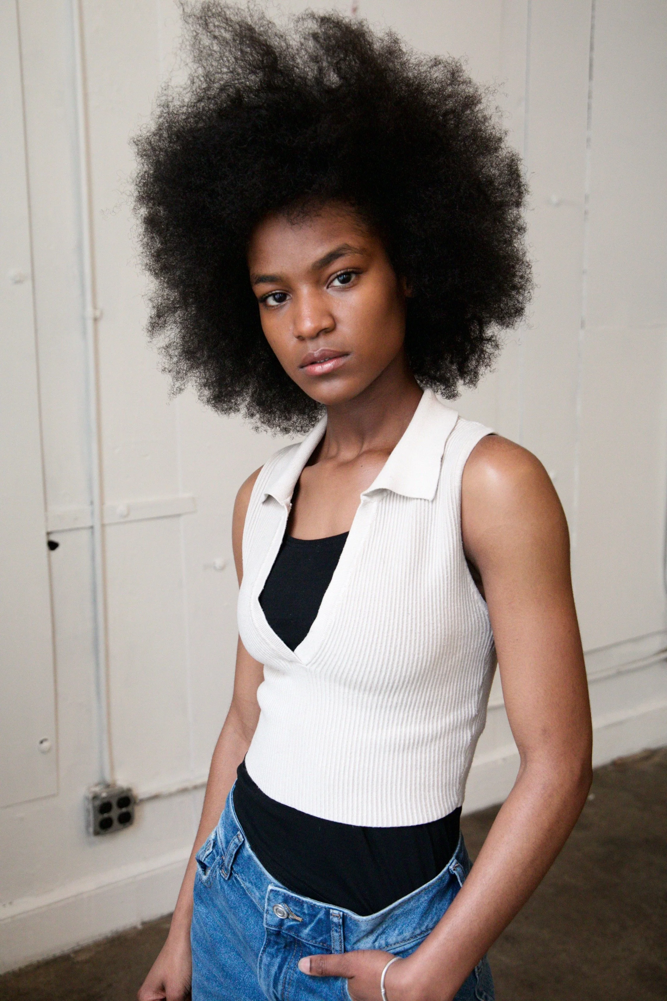 A woman with natural afro hair standing in front of a plain wall. She is wearing a sleeveless beige ribbed top layered over a black shirt, and blue jeans. Her expression is serious, and she has a silver bracelet on her left wrist.
