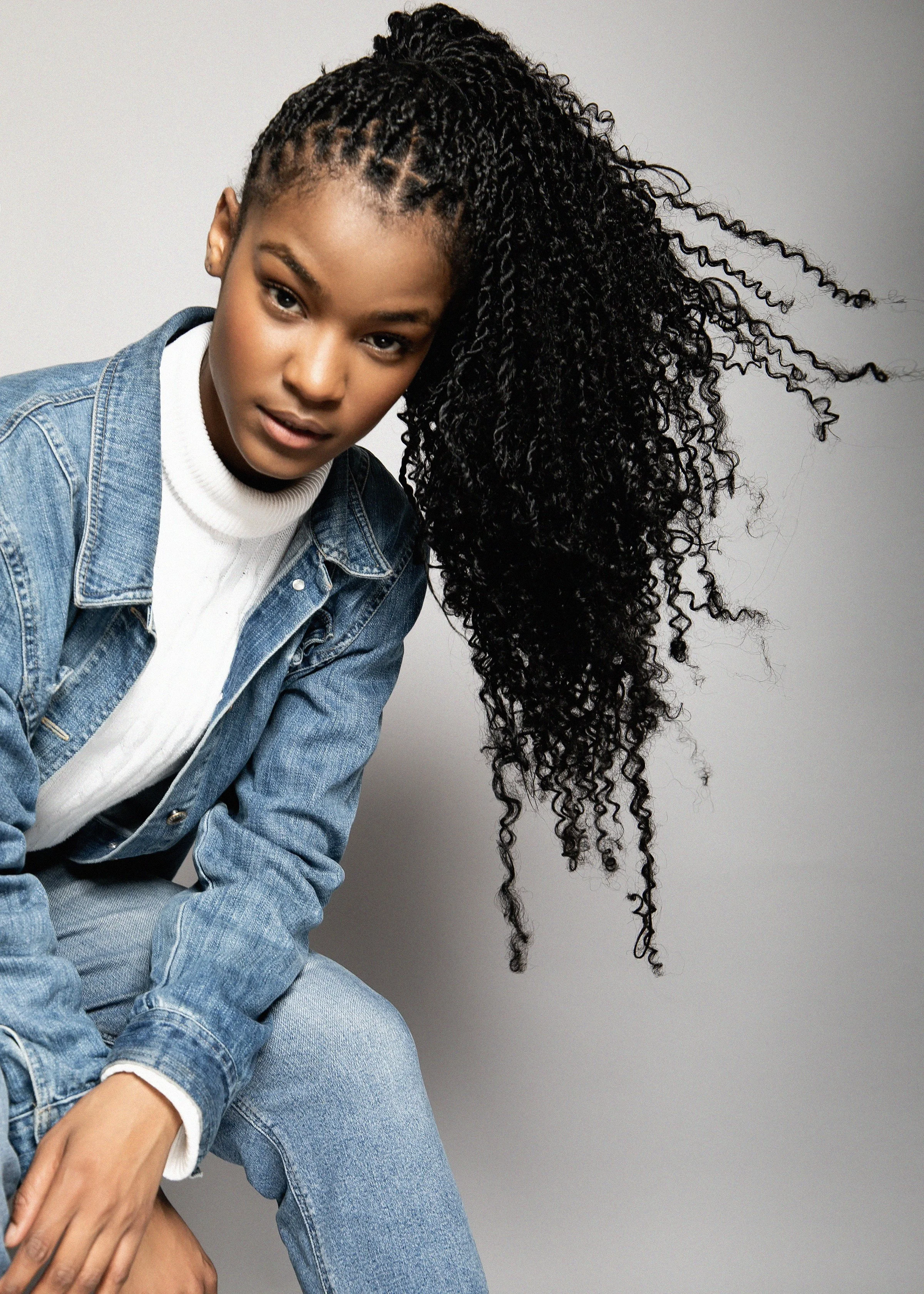 A young woman with dark skin and curly hair, wearing a denim jacket and white turtleneck, poses against a plain gray background.