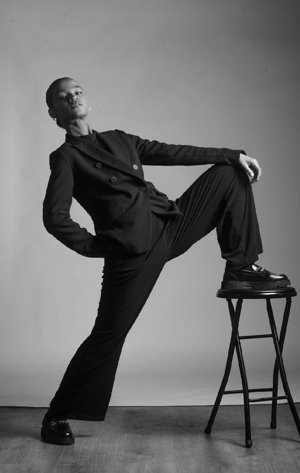 A black and white photo of a woman in a stylish dark outfit, standing with one foot on a chair, posing confidently against a neutral background.