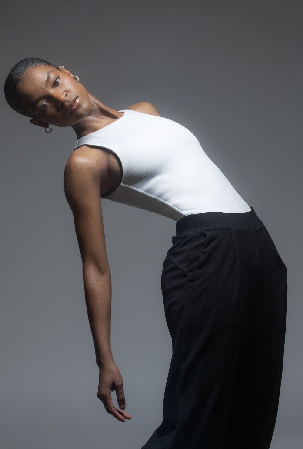 A woman in a white sleeveless top and black pants leaning to the side against a gray backdrop.