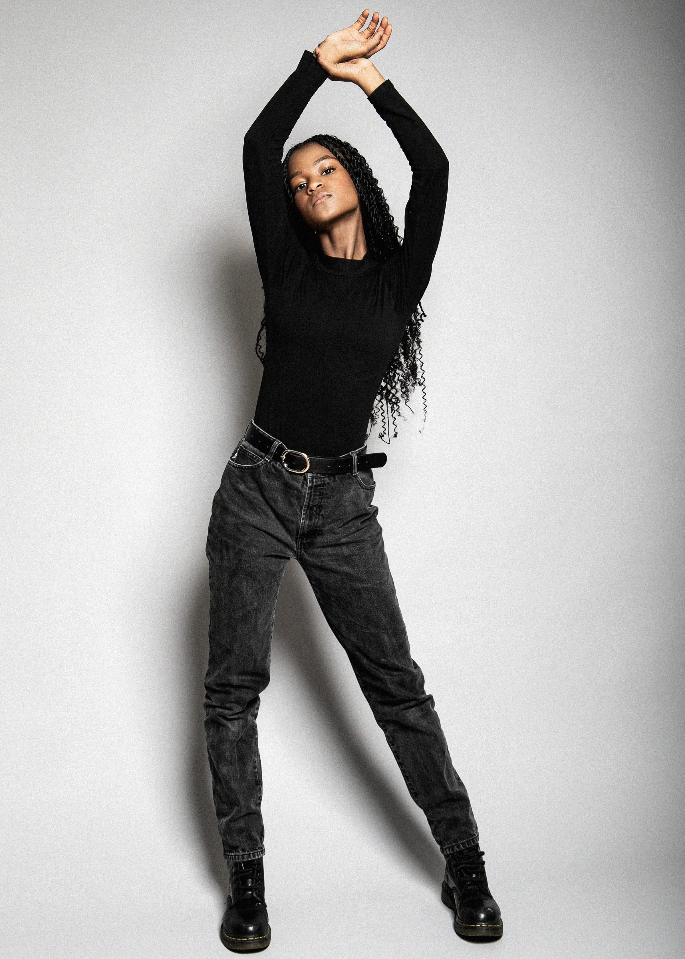 A woman with long black curly hair wearing a black long-sleeve shirt, black high-waisted jeans, a black belt, and black boots posing with her arms raised above her head against a plain light grey background.