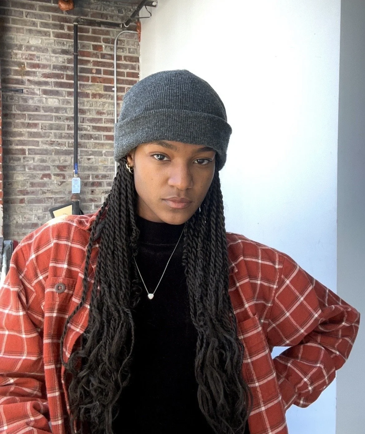 A young woman with long braided hair wearing a gray beanie, a black top, a red plaid shirt, and a delicate necklace. She is standing indoors with a brick wall and some industrial piping in the background, looking directly at the camera with a serious expression.