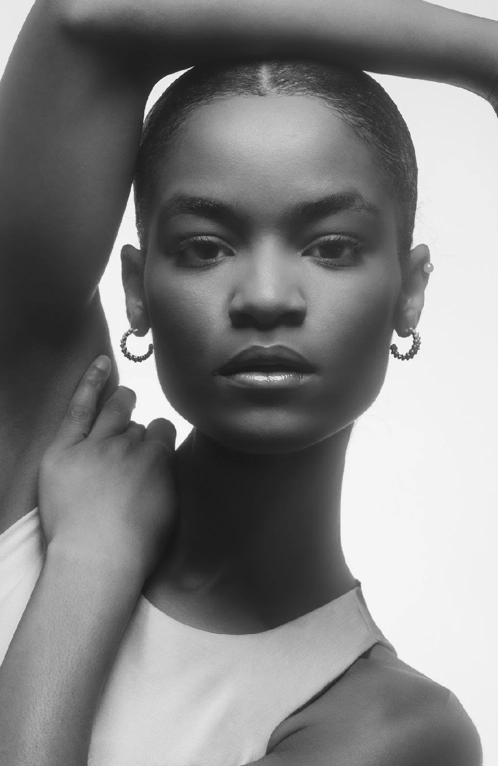 Black and white portrait of a woman with her arm raised above her head, wearing hoop earrings and a sleeveless top, with a confident expression.