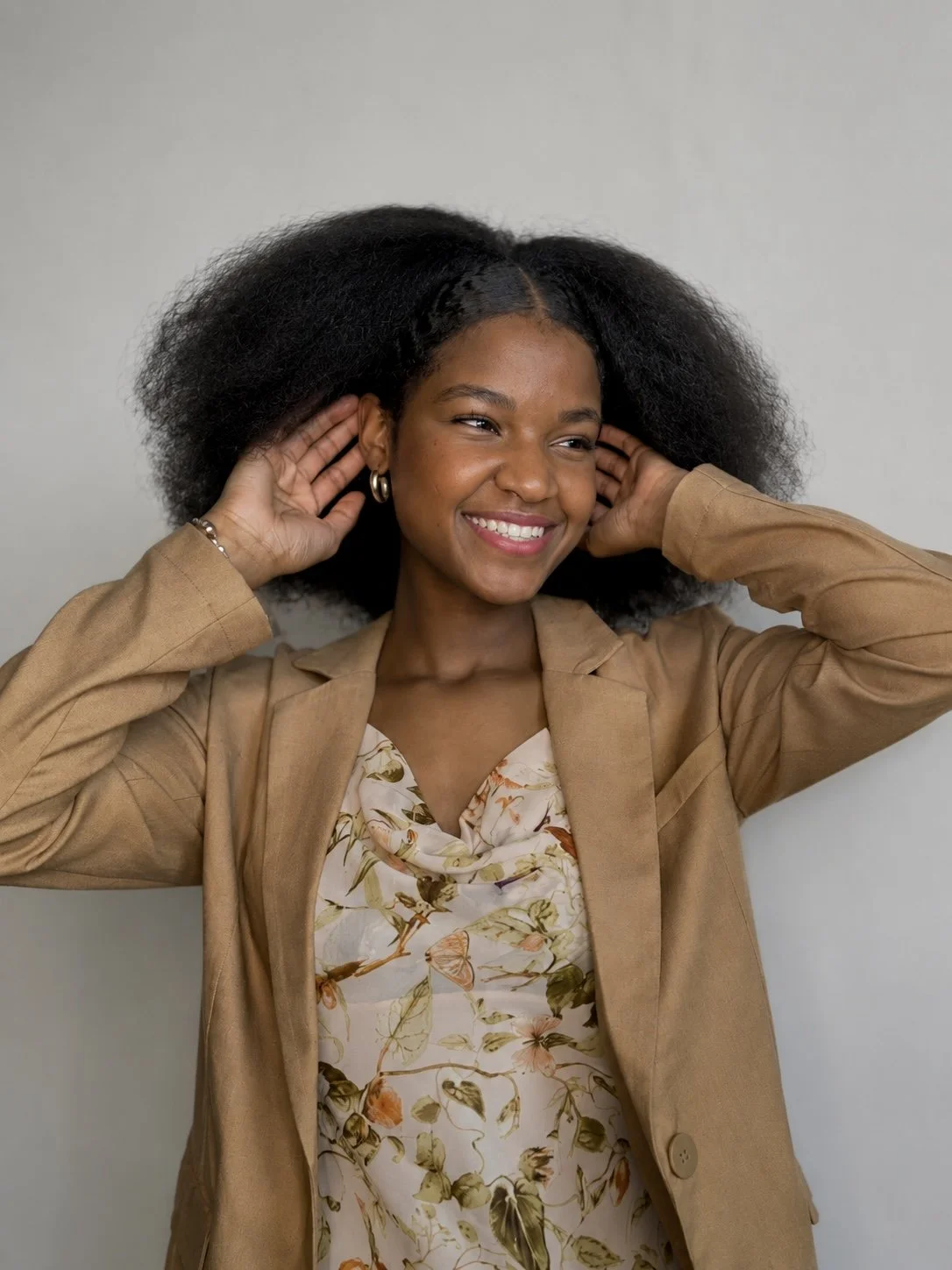 A smiling woman with natural hair, wearing a floral blouse and a tan blazer, standing against a plain gray background.