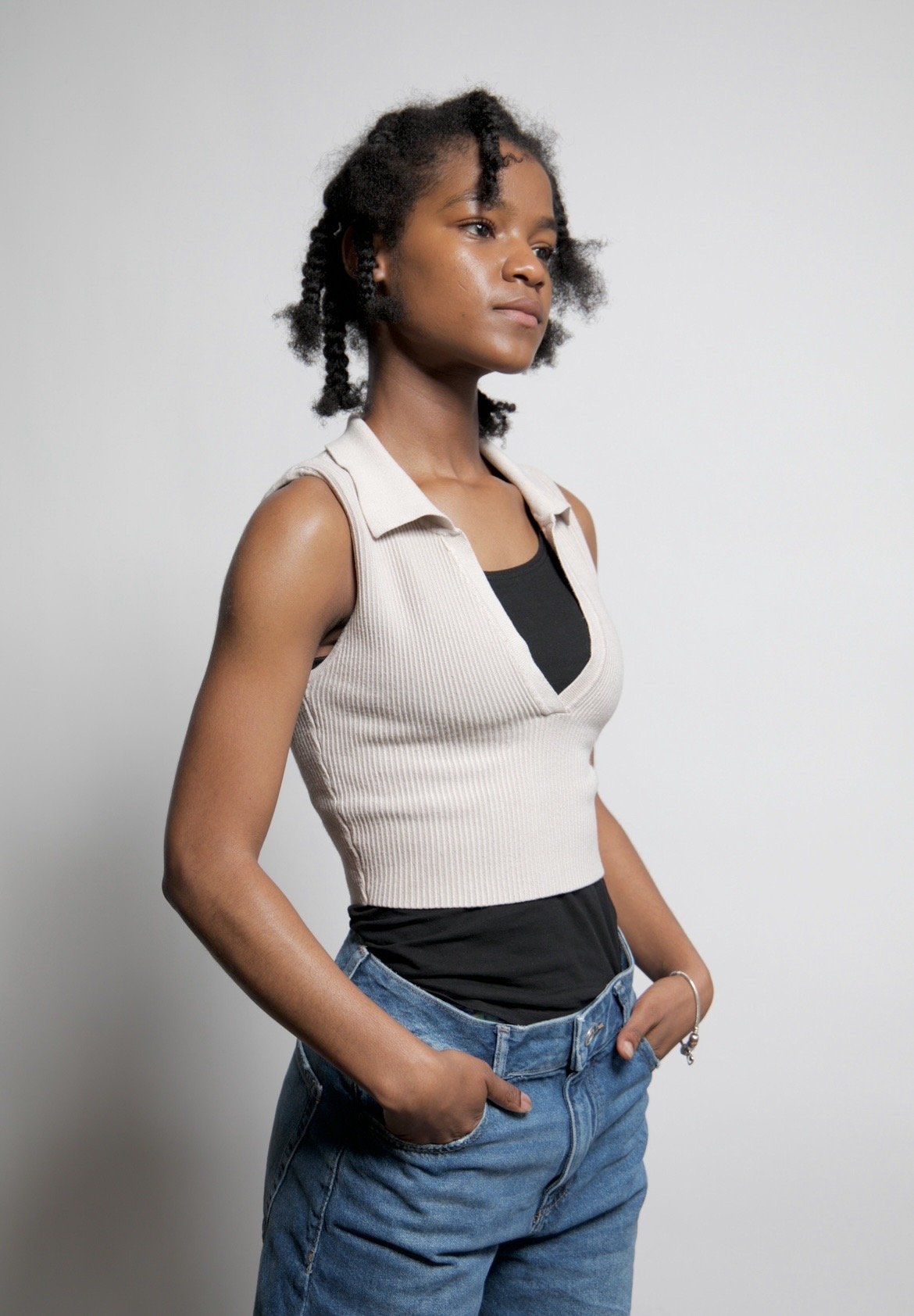 A young woman standing against a plain light gray background, with short, natural curly hair, wearing a sleeveless beige vest over a black top, and blue jeans.