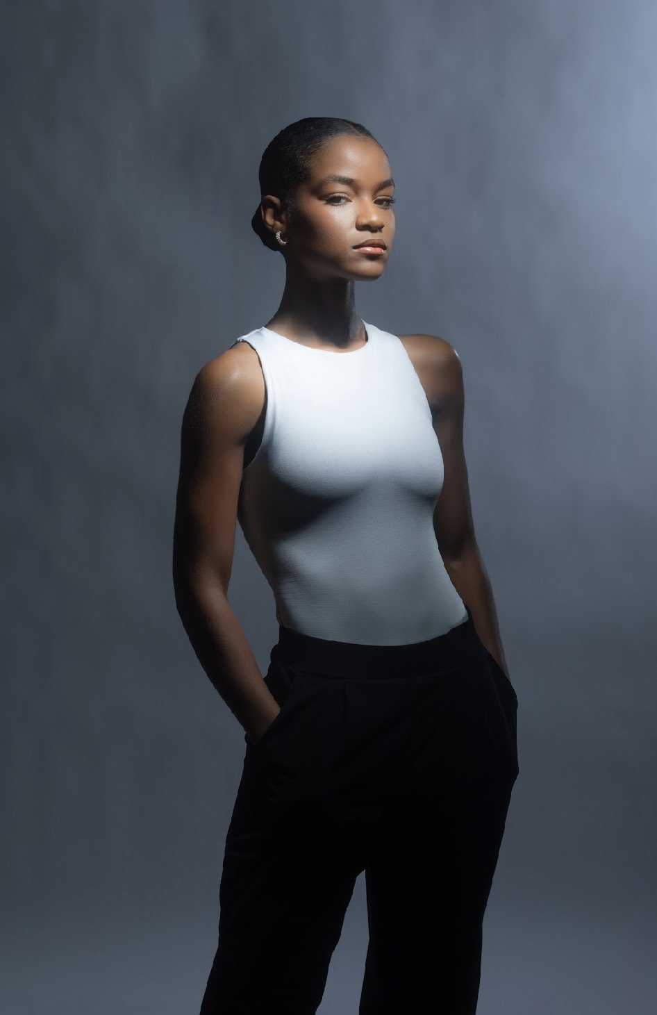 A confident woman with short hair in a sleeveless white top and black pants, posing against a gradient gray background.