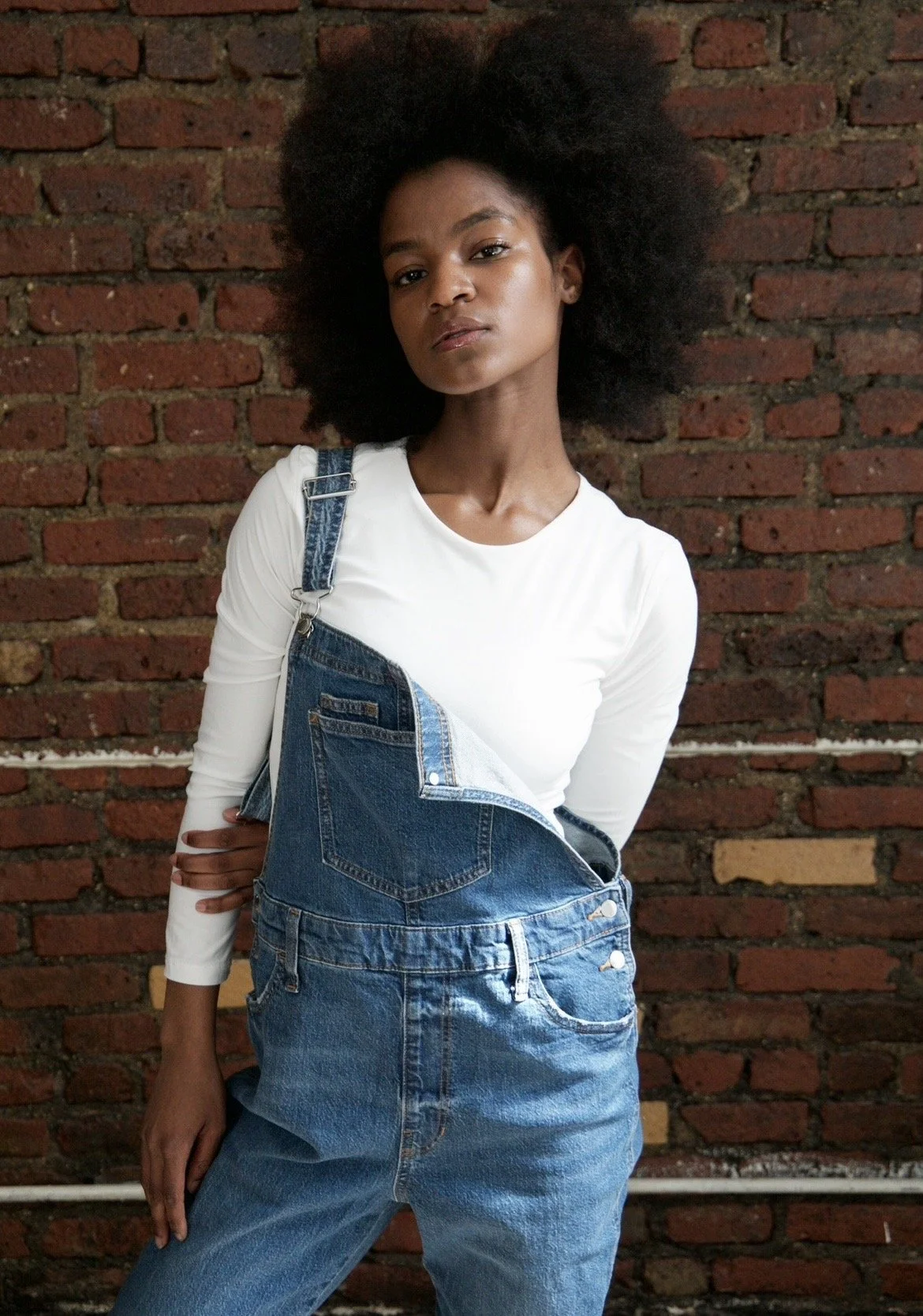 A woman with natural curly hair wearing a white long-sleeve shirt and denim overalls standing against a red brick wall.