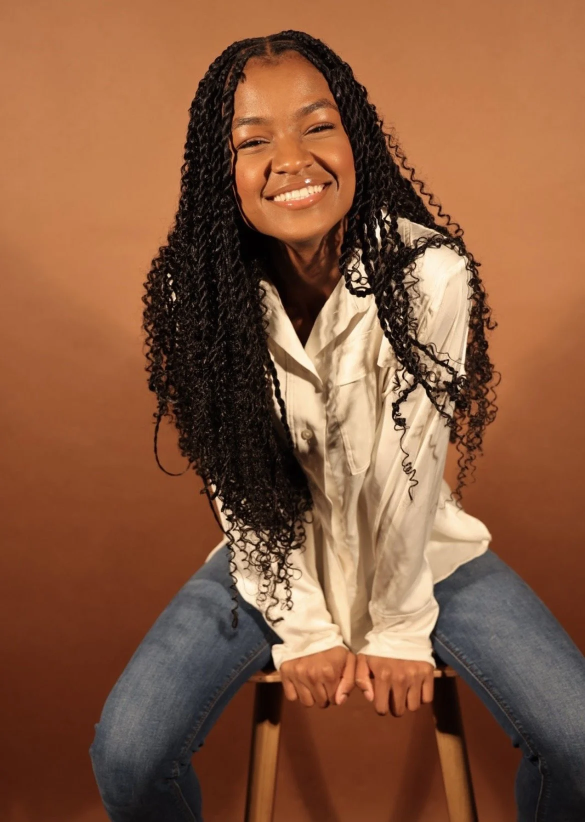 A woman with long, curly black hair, smiling and sitting on a stool against a brown background, wearing a white shirt and blue jeans.