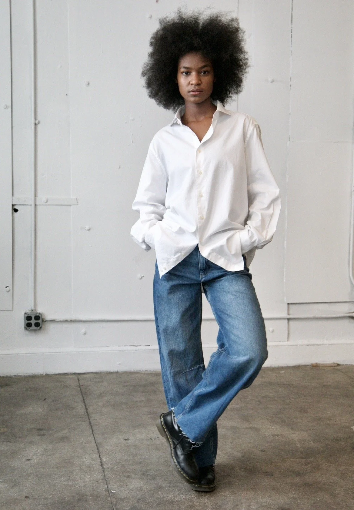 A woman with natural curly hair wearing a white shirt, blue jeans, and black boots standing with hands in pockets against a plain white wall.