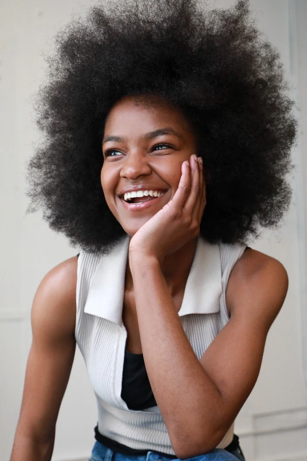 A woman with a big afro hairstyle smiling and touching her face with her hand.