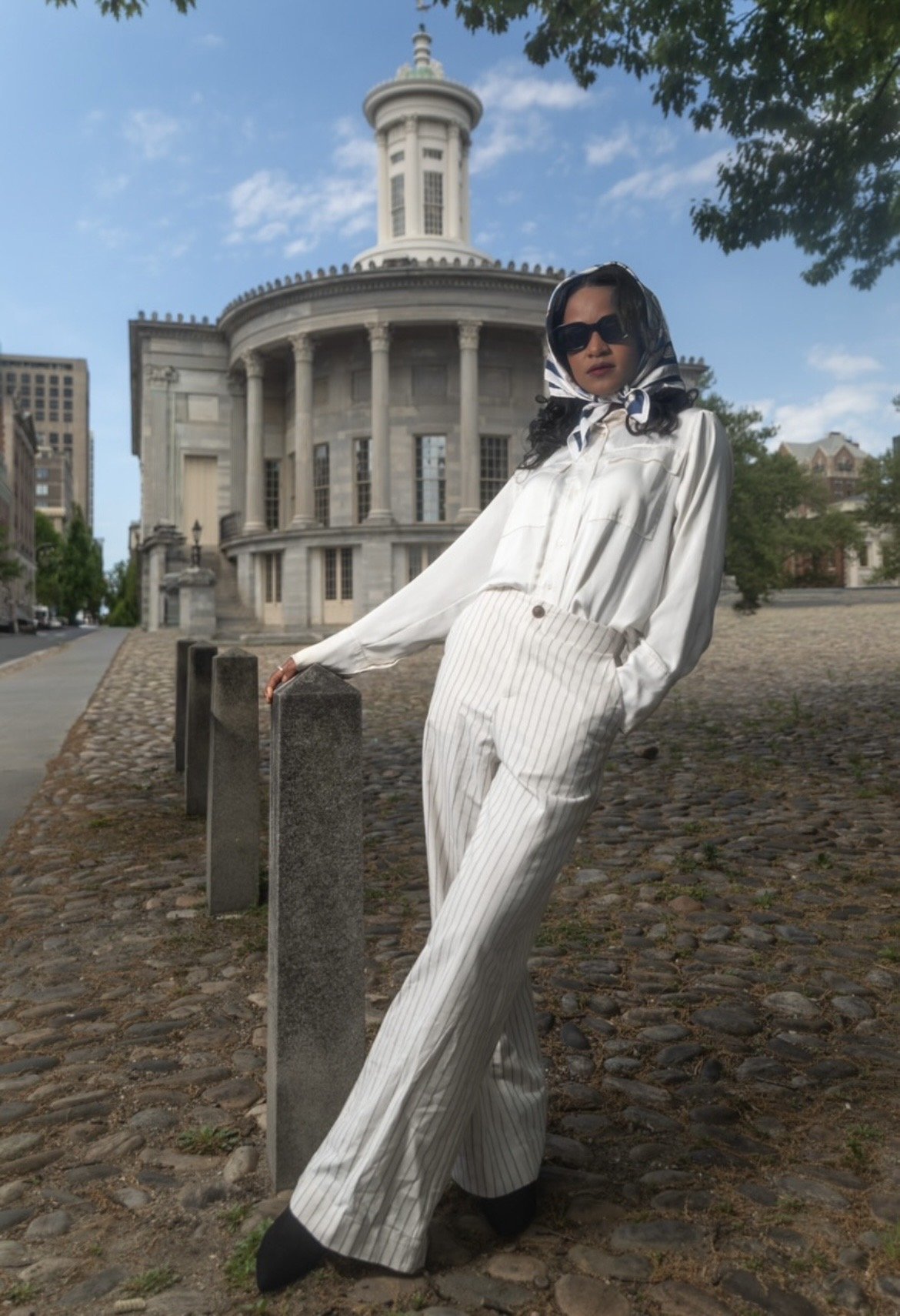 Woman in striped white and beige pants, white silk shirt, black sunglasses, and a patterned headscarf posing outdoors in front of a historic building with columns, under a partly cloudy sky.