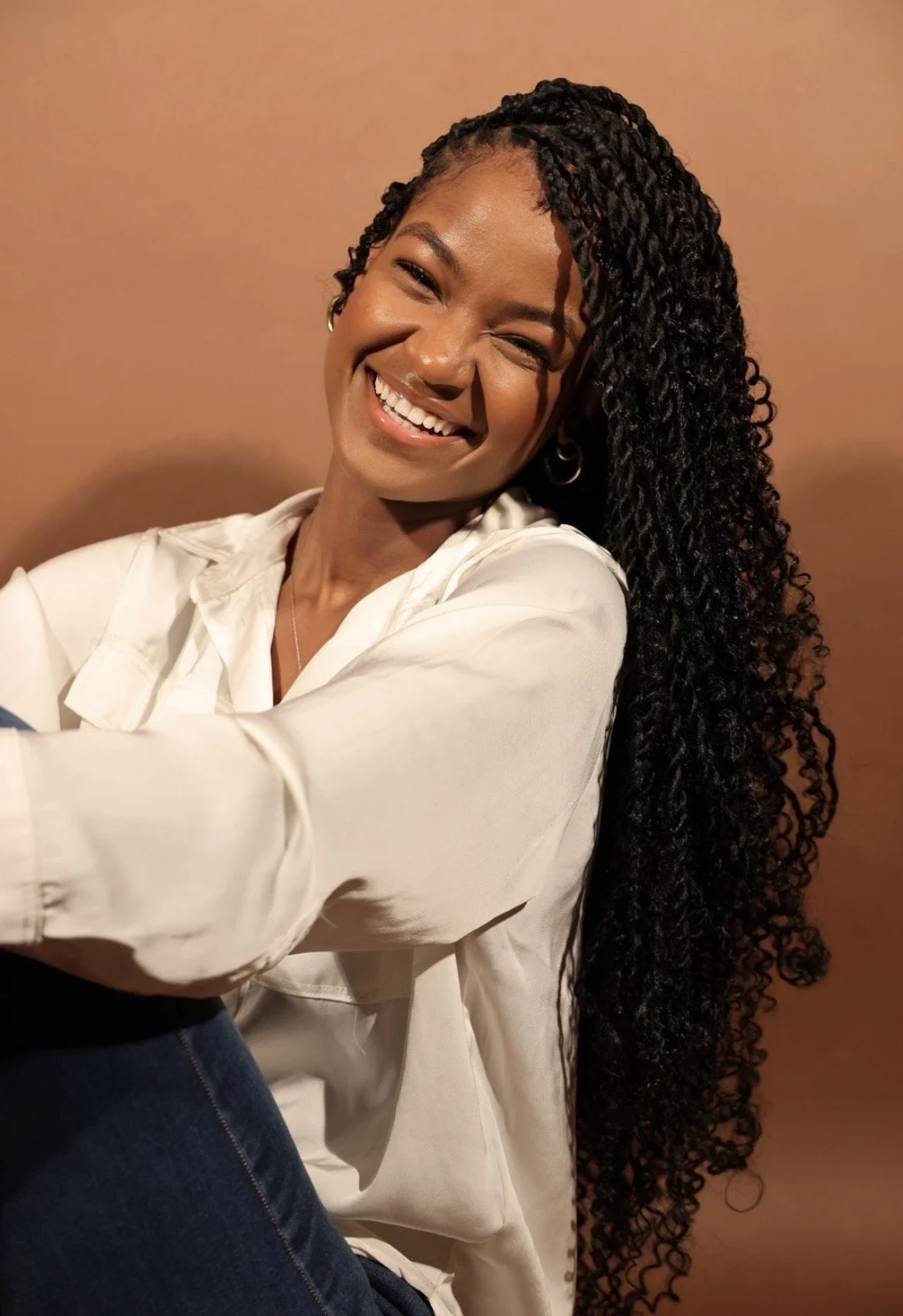 A woman with dark, curly hair in a twist style, smiling and winking at the camera, wearing a white shirt against a plain brown background.